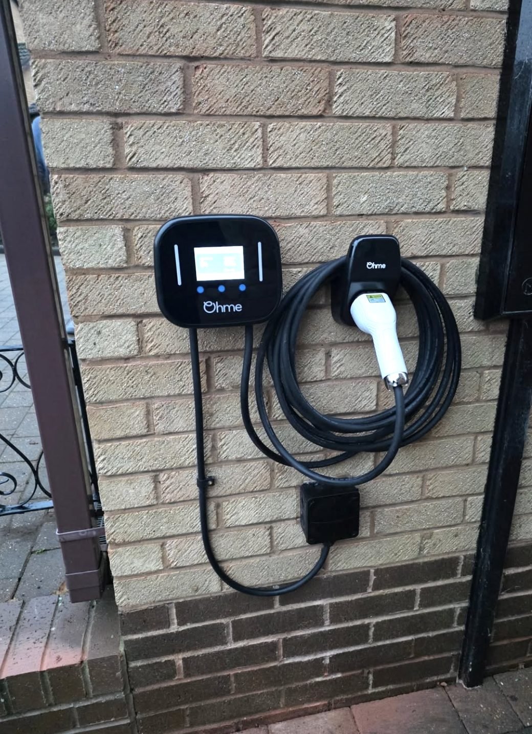 Ohme tethered EV charger with coiled cable beside a driveway gate